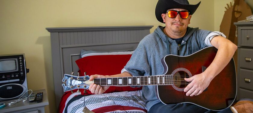 A man wearing sunglasses and a cowboy hat sitting on his bed and playing guitar
