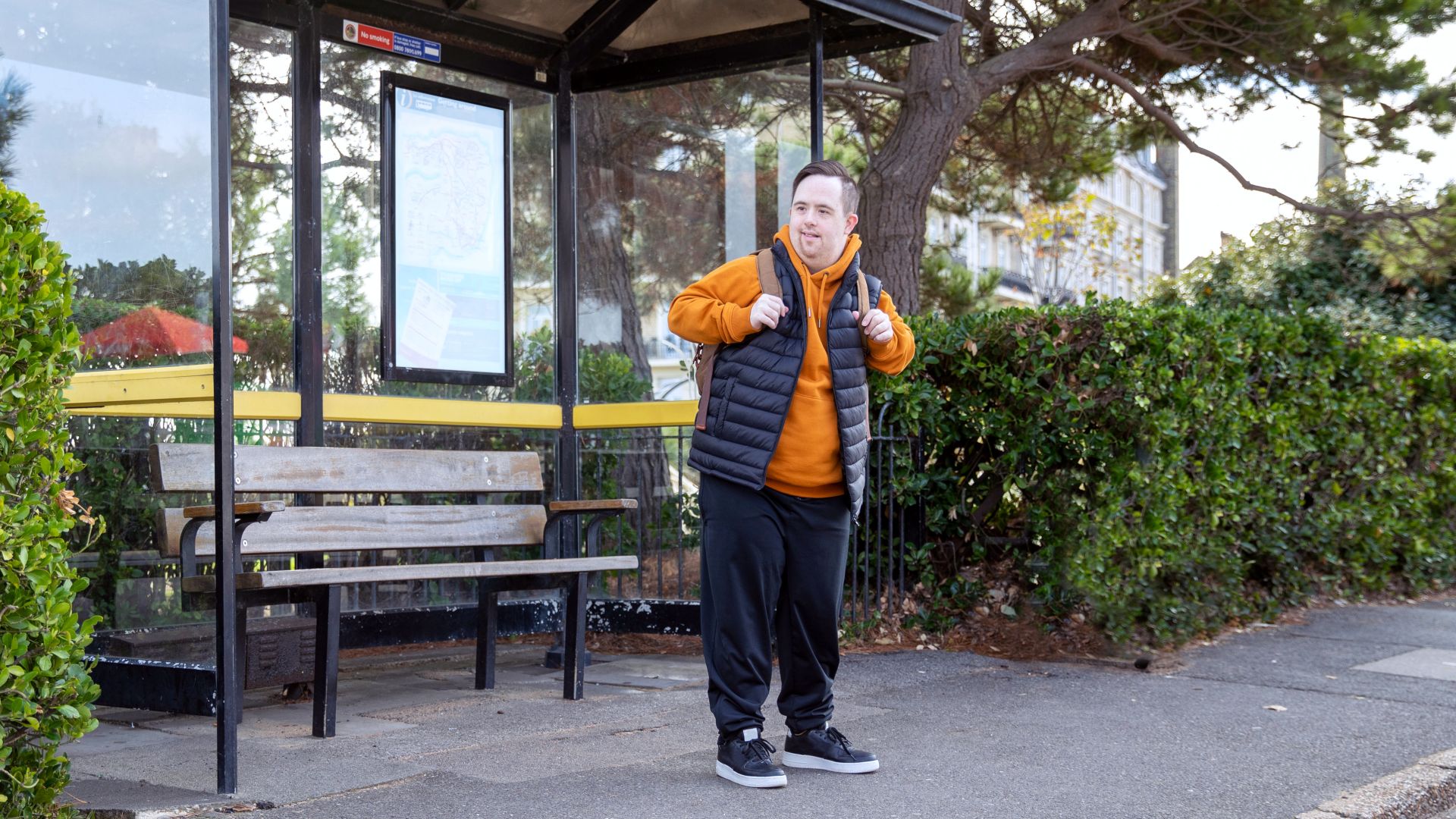 A man wearing an orange hoodie and a backpack eagerly waiting at a bus stop.