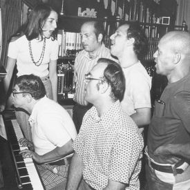 Black and white photo of group of adults singing and playing piano.