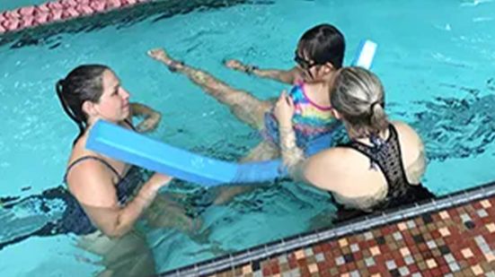 Two women teaching a young child to swim with a pool noodle.