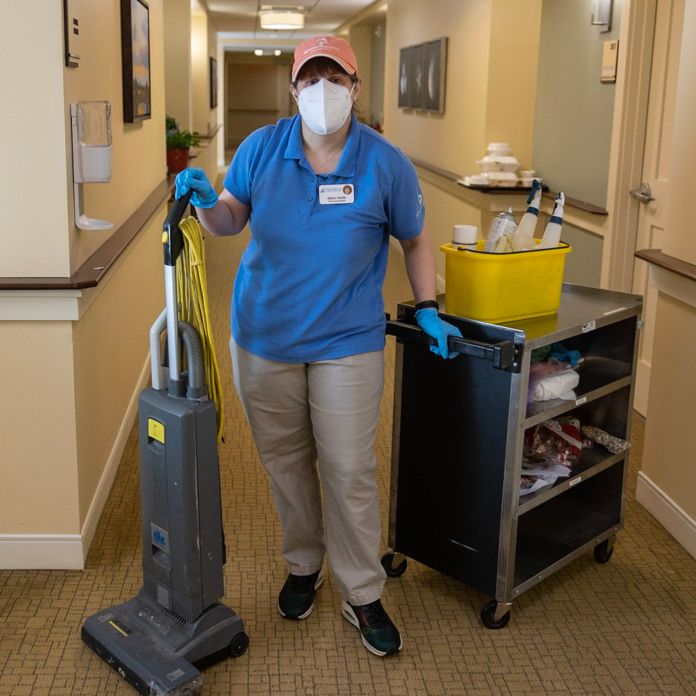 Janitorial worker in a pink hat posing next to a vacuum and a cart of cleaning equipment.