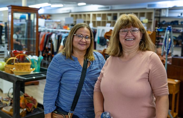 Two adult females with glasses standing and smiling inside a thrift store.