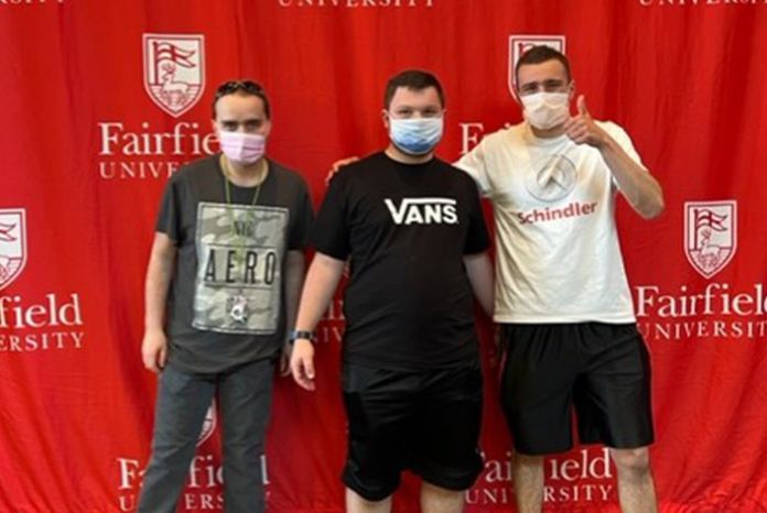 Three young adults posing in front of a red Fairfield University backdrop.
