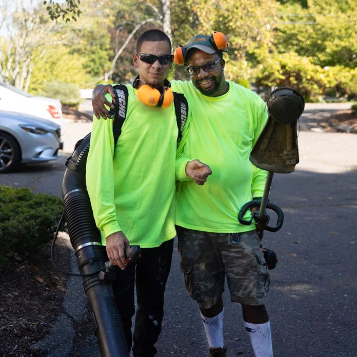Two men holding a weed whacker and leaf blower smiling.