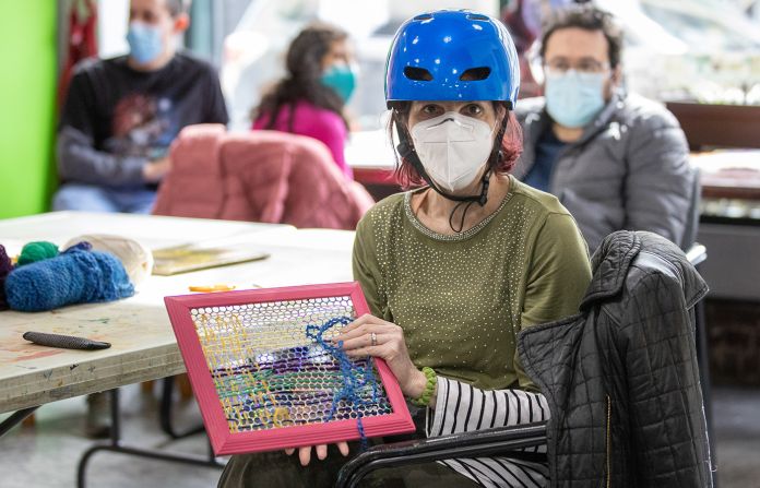 Woman with a mask and blue helmet sitting and holding artwork made of yarn.