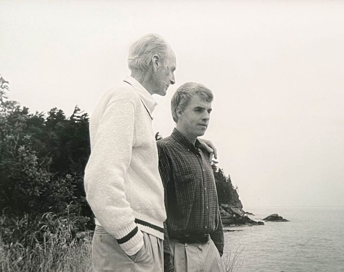 Two men overlooking the water with their arms around each other's shoulders. 