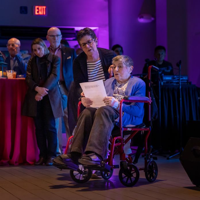 Woman in a wheelchair holding a piece of paper speaking to a crowd at an event.
