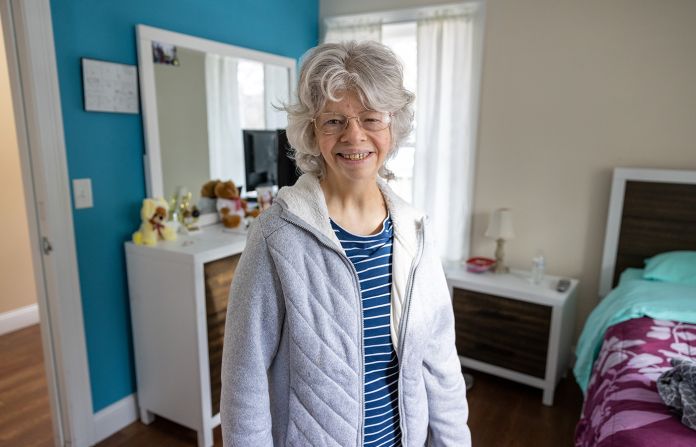 Woman with grey hair standing and smiling in her home bedroom. 