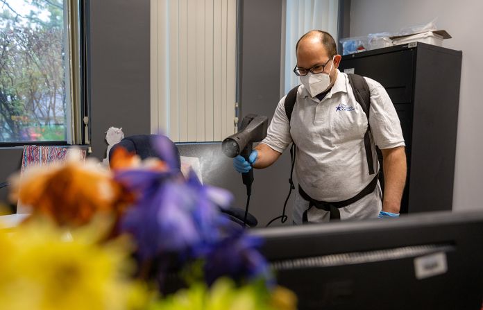 Man in a mask spraying a disinfectant mist in an office with colorful flowers.